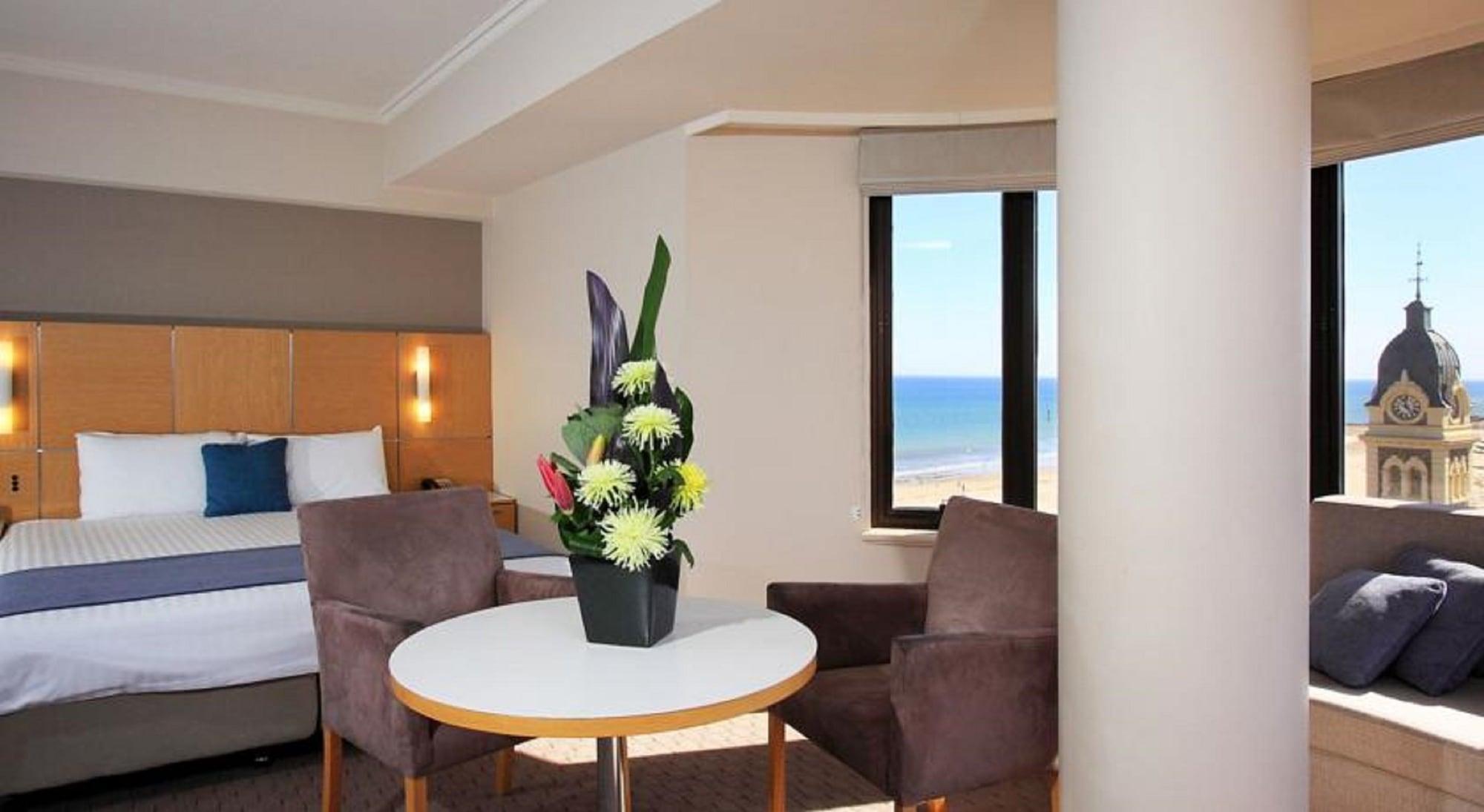 מלון גלנלג Stamford Grand Adelaide מראה חיצוני תמונה The photo shows a cozy hotel room with a comfortable seating arrangement. There are two brown chairs positioned around a round table, which features a decorative flower arrangement in a black vase. The background includes a window that offers a view 