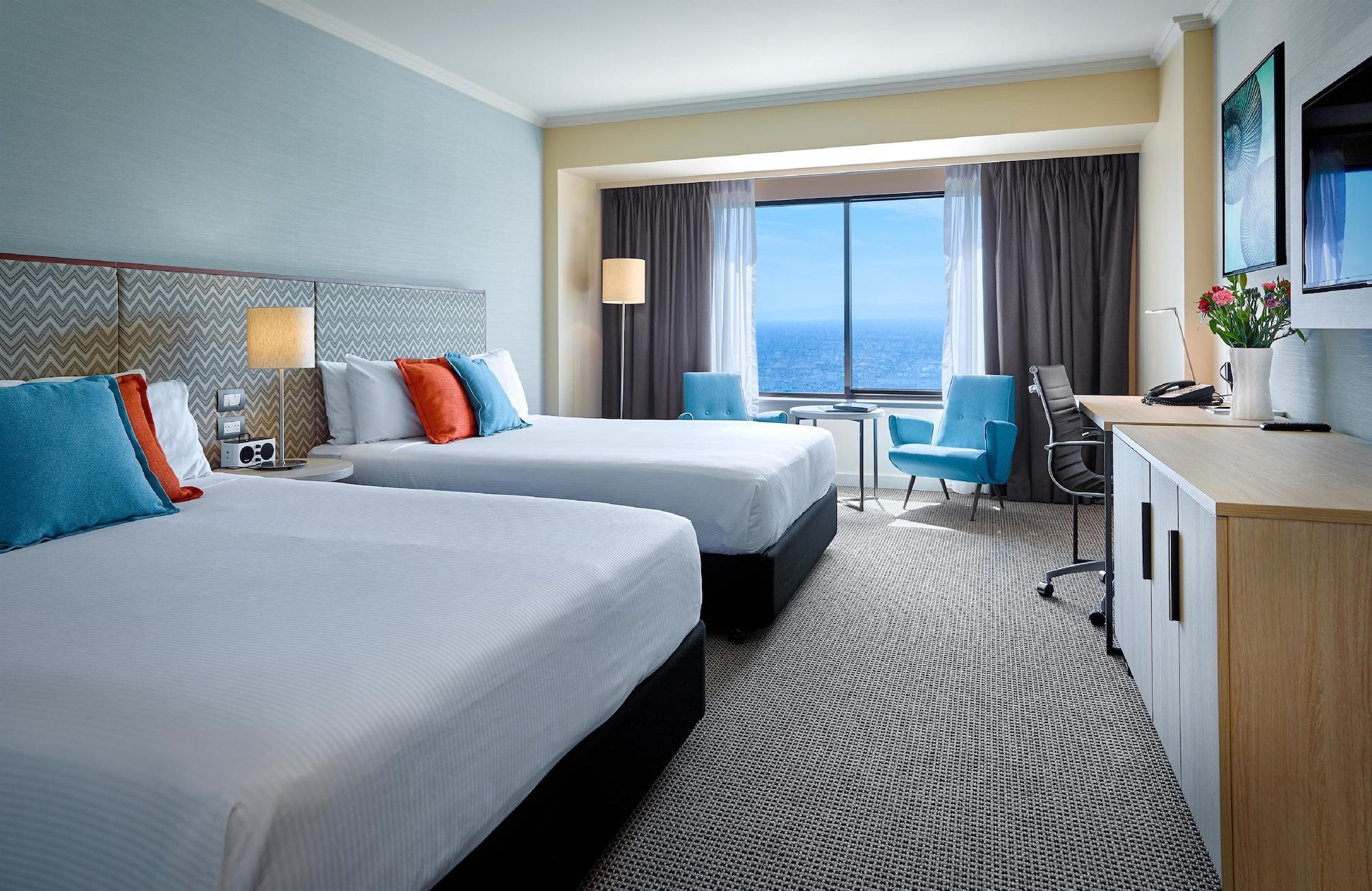 מלון גלנלג Stamford Grand Adelaide מראה חיצוני תמונה The photo shows a well-decorated hotel room featuring two queen-sized beds with white linens and colorful accent pillows. There is a large window with curtains that offers a view of the ocean. A small seating area with a blue chair and a matching des
