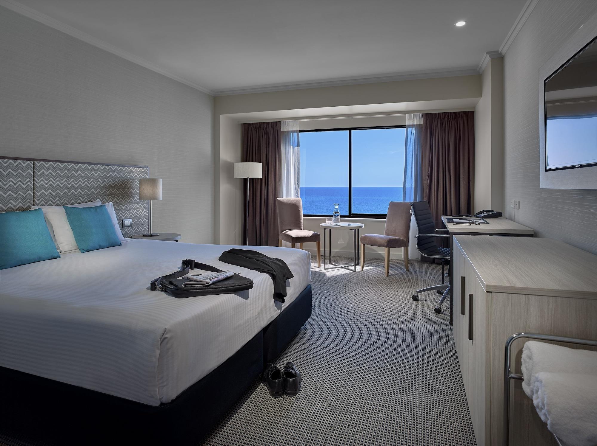 מלון גלנלג Stamford Grand Adelaide מראה חיצוני תמונה The photo shows a modern hotel room featuring a queen-sized bed with white linens and two aqua blue decorative pillows. There are bedside lamps and a large window with a view of the ocean. The room also has a small seating area with two chairs and a 