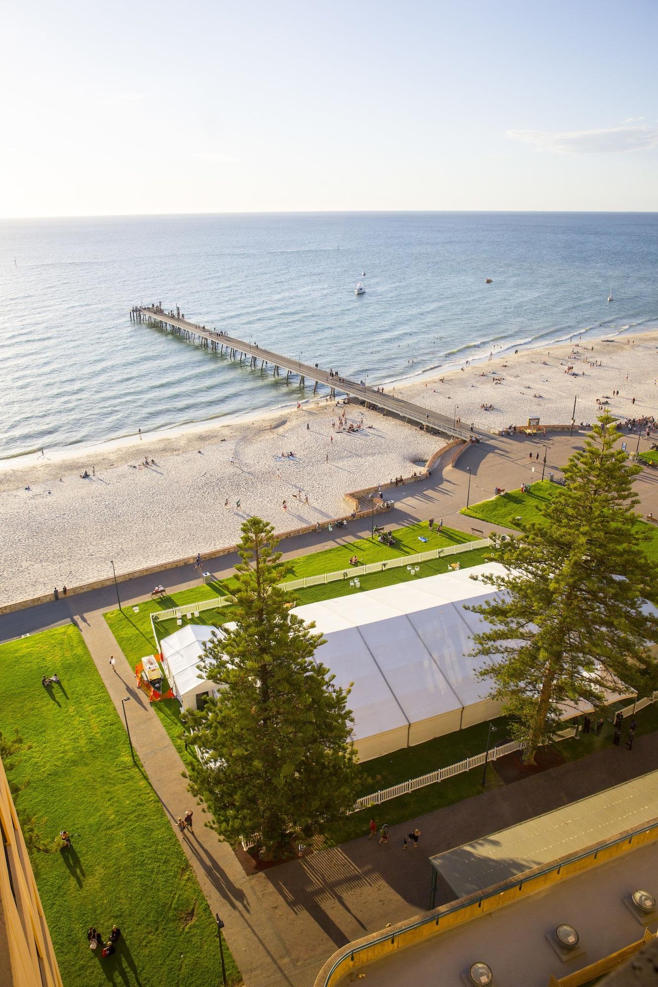 מלון גלנלג Stamford Grand Adelaide מראה חיצוני תמונה The photo shows a scenic view of a beach with a long pier extending into the water. The beach is sandy, and there are people enjoying the sun and walking along the shore. In the foreground, there is a large white tent set up on a grassy area, likely 