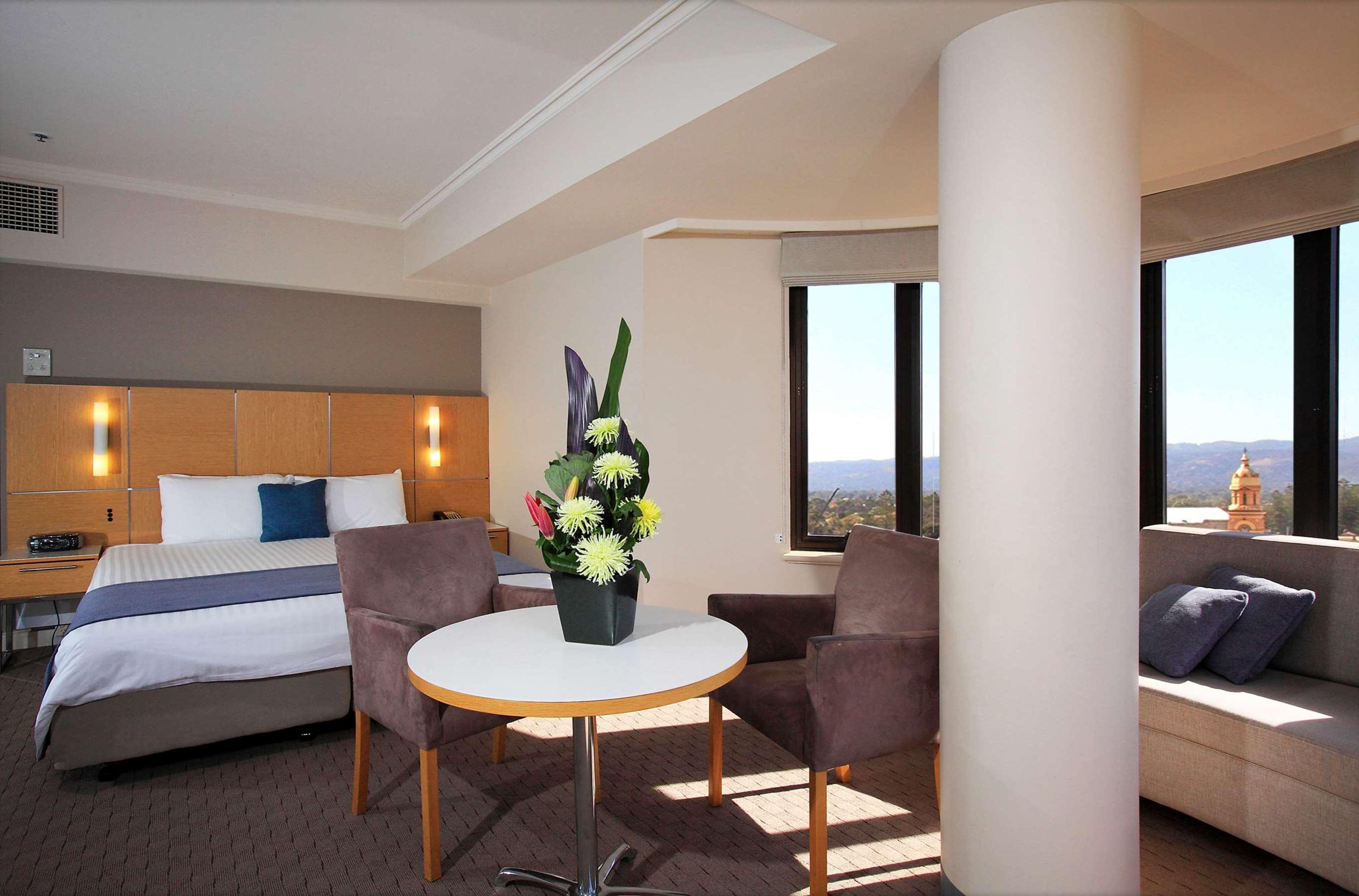 מלון גלנלג Stamford Grand Adelaide מראה חיצוני תמונה The photo shows a hotel room with a modern design. On one side, there is a plush bed with white and blue bedding. In the center of the room, there's a small round table with two gray upholstered chairs around it. A vase of flowers sits on the table, 