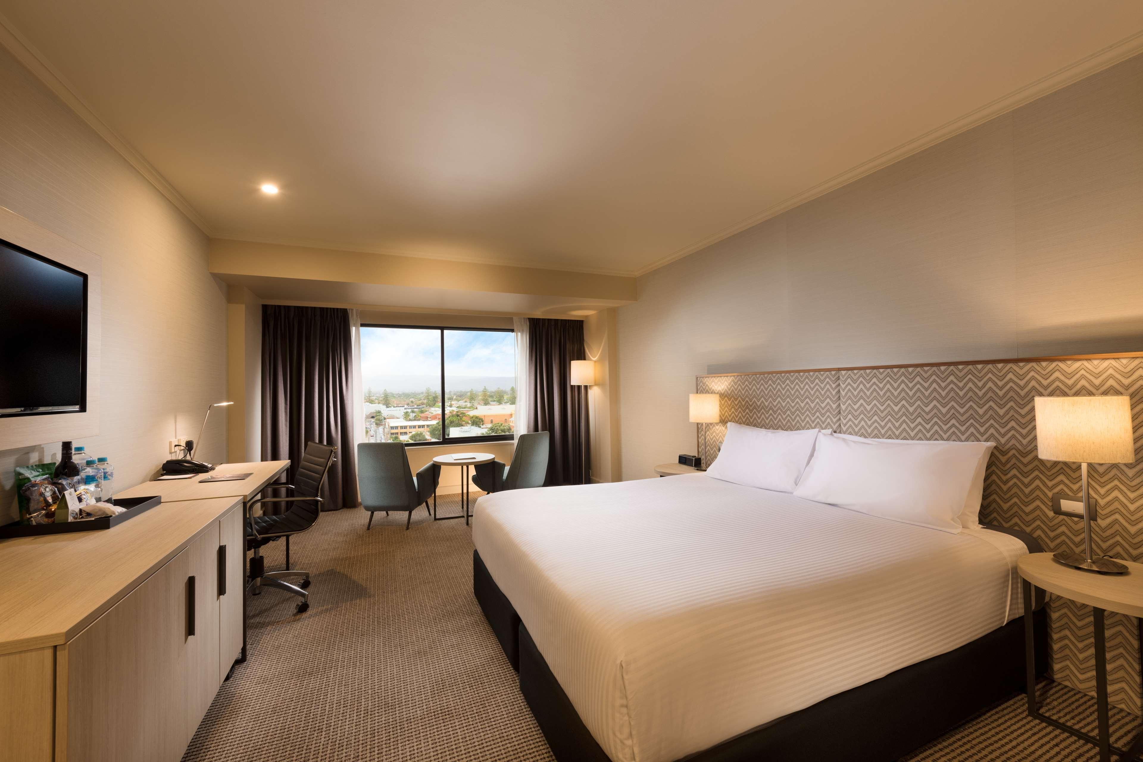 מלון גלנלג Stamford Grand Adelaide מראה חיצוני תמונה The photo shows a modern hotel room. It features a large bed with white linens, a desk with a chair, and a flat-screen TV mounted on the wall. There are two armchairs and a small table near a large window that lets in natural light, providing a view 