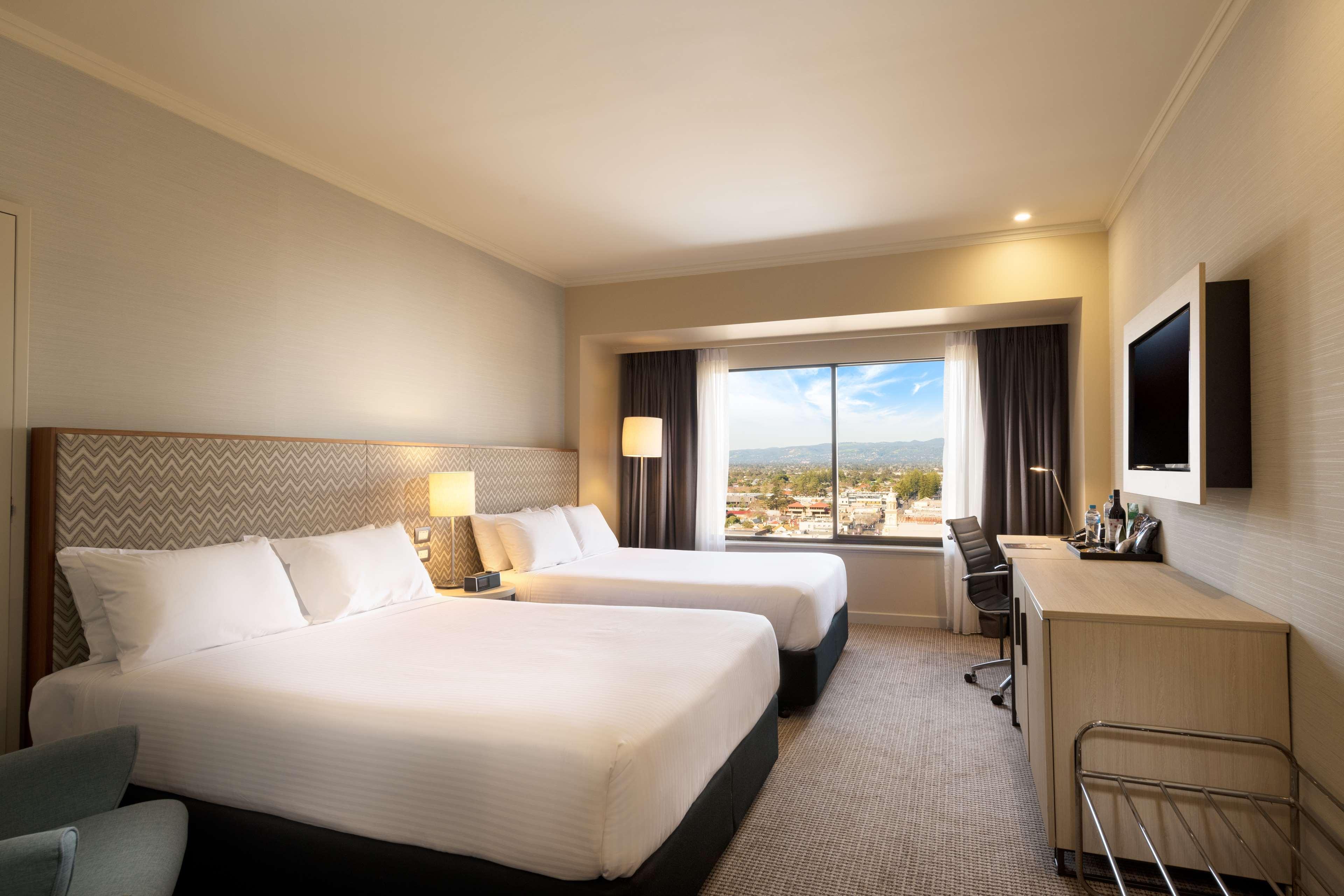 מלון גלנלג Stamford Grand Adelaide מראה חיצוני תמונה The photo shows a modern hotel room featuring two beds with white linens. There are bedside tables with lamps next to each bed. A large window provides natural light and offers a view of the outside, with curtains on either side. The room has a neutr