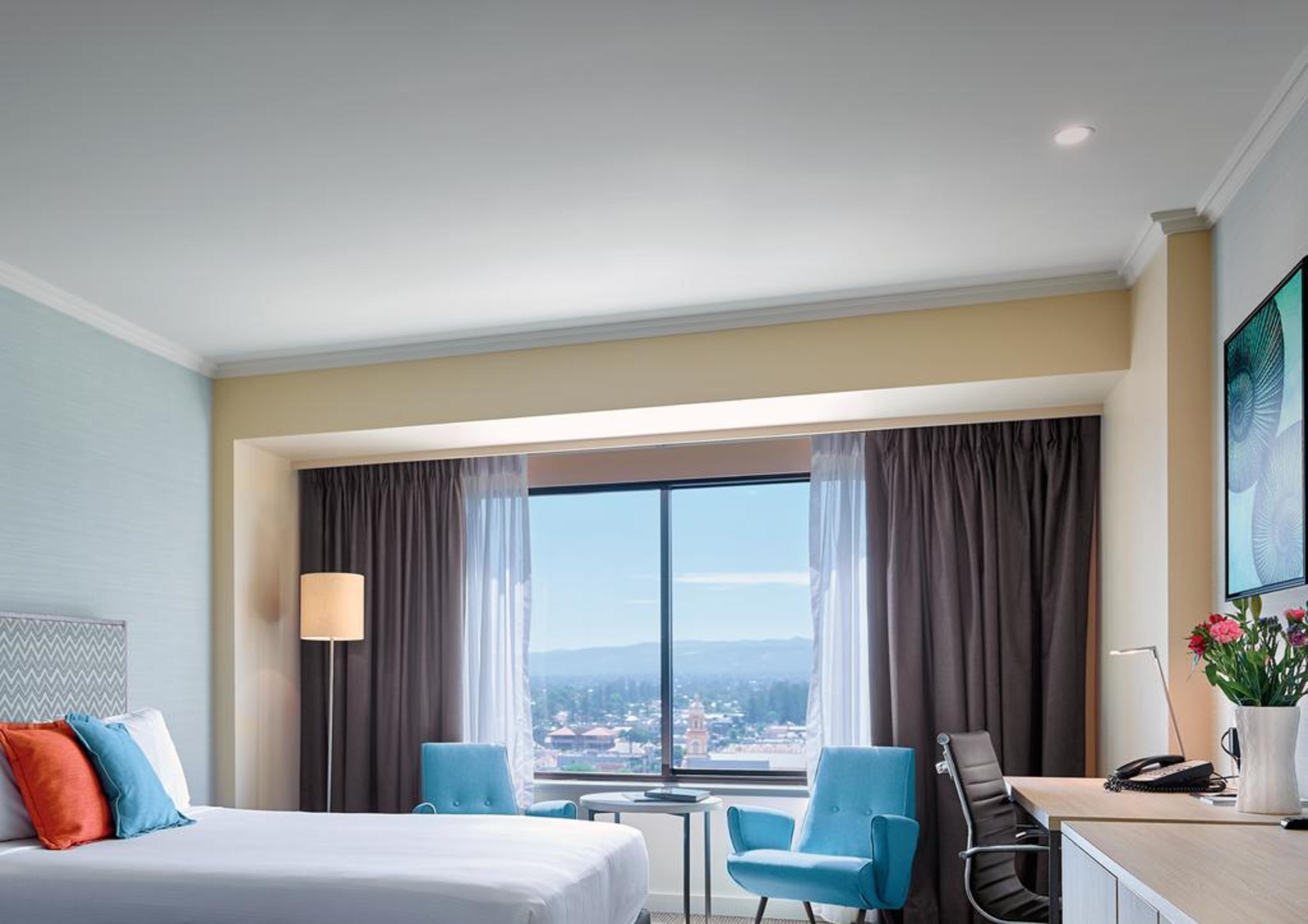 מלון גלנלג Stamford Grand Adelaide מראה חיצוני תמונה The photo shows a modern hotel room featuring a queen-sized bed with white linens and colorful decorative pillows. There are two blue chairs beside a small table positioned near a large window that provides a view of the outside, with sheer and dark 