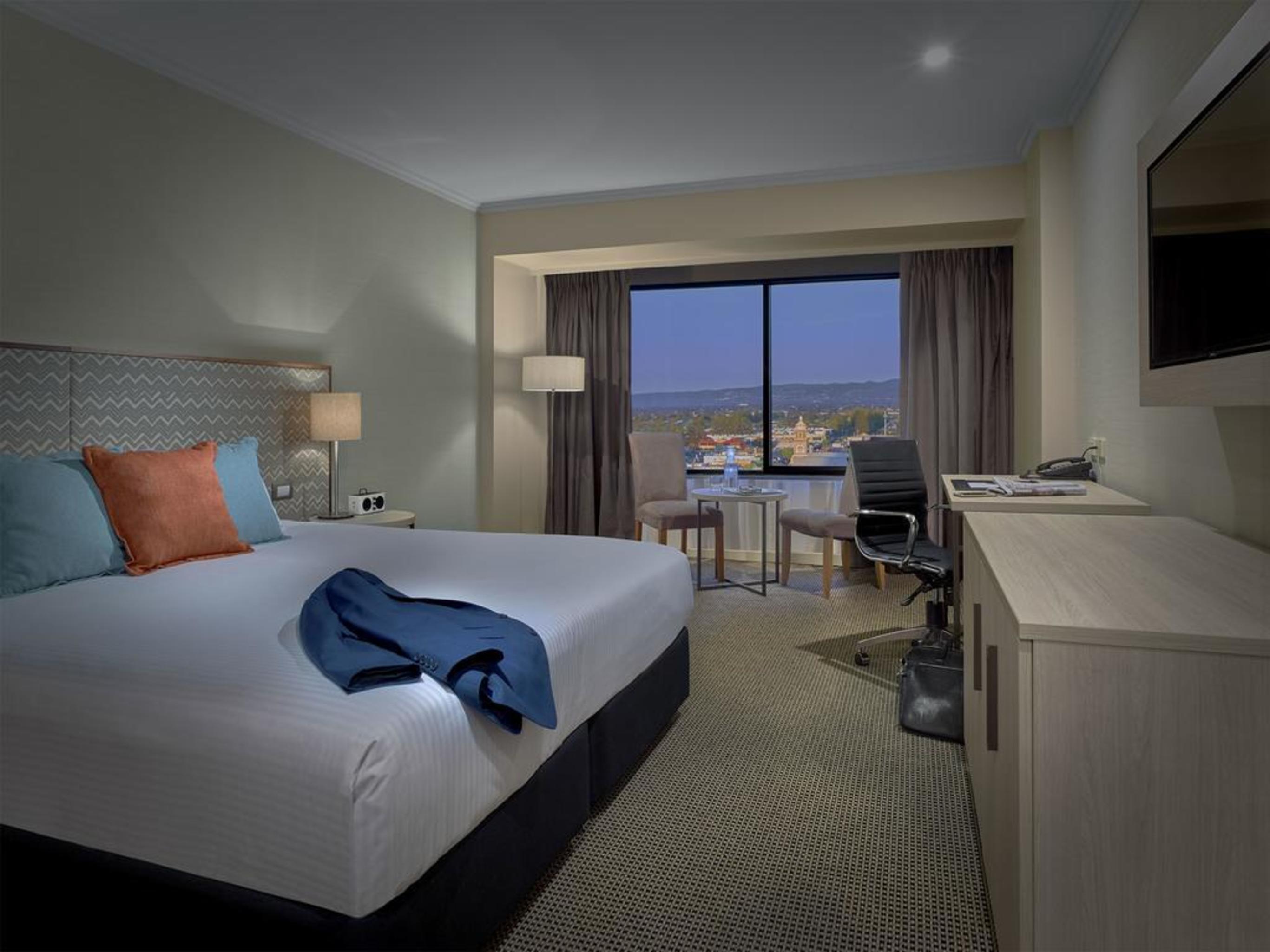 מלון גלנלג Stamford Grand Adelaide מראה חיצוני תמונה The photo shows a modern hotel room. It features a large double bed with white bedding and decorative pillows in orange and blue. There's a nightstand with lamps on either side of the bed. A large window overlooks a view outside, and there's a desk a