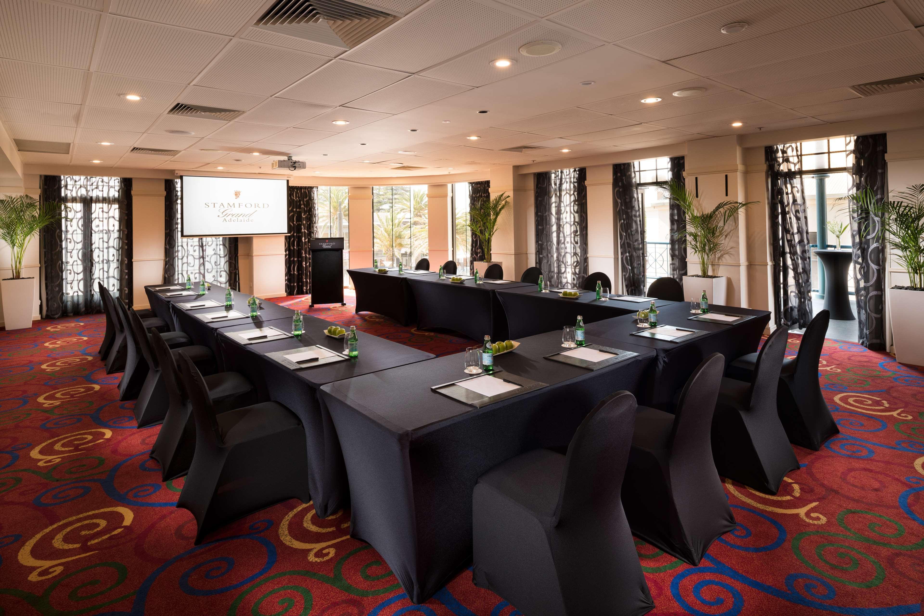 מלון גלנלג Stamford Grand Adelaide מראה חיצוני תמונה The photo shows a conference room set up for a meeting or presentation. There are several tables arranged in a U-shape, covered with black tablecloths. Each table has a notepad and a small bottle of water on it. The chairs around the tables are also 