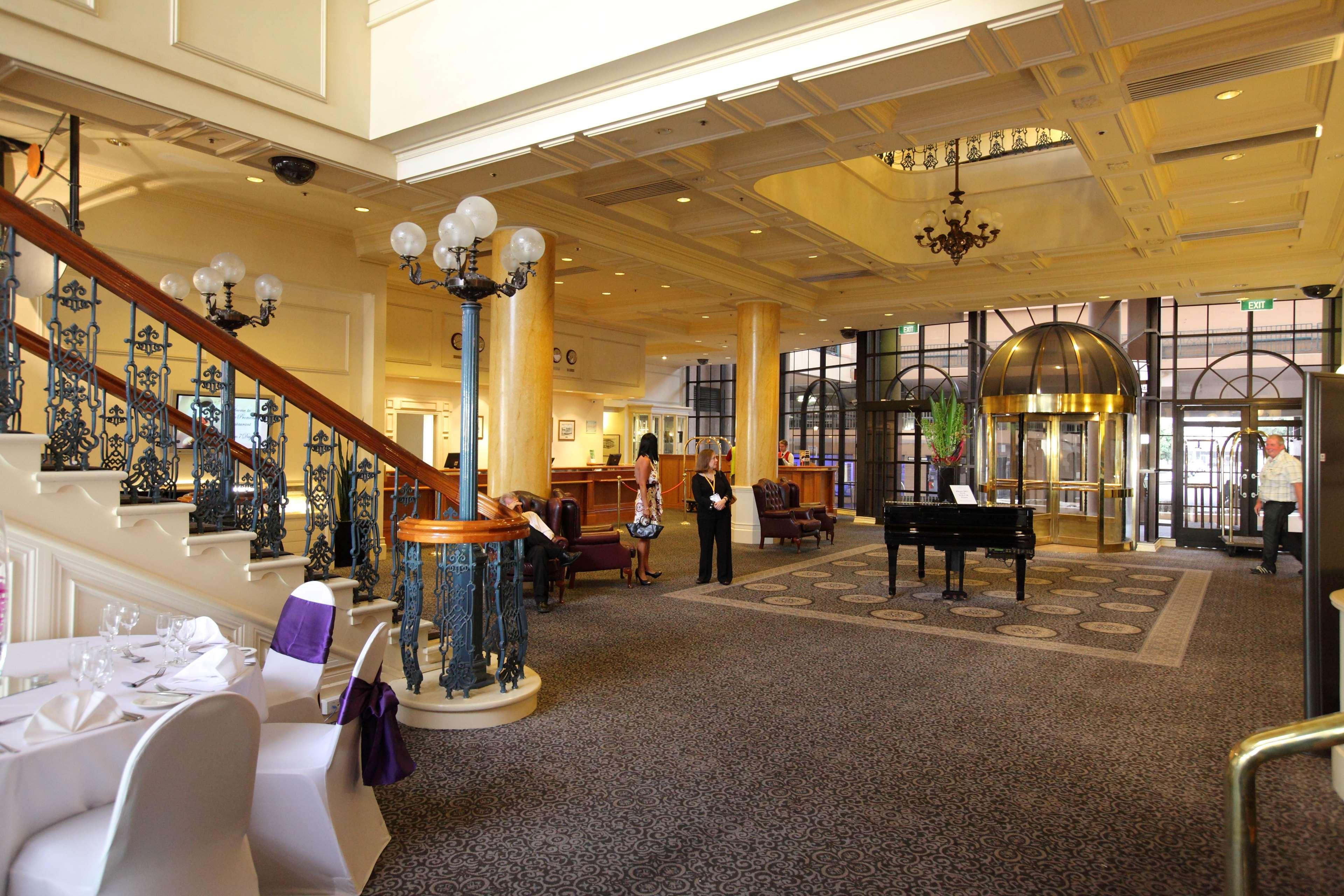 מלון גלנלג Stamford Grand Adelaide מראה פנימי תמונה The photo shows a luxurious interior of a hotel or a restaurant lobby. There is an elegant staircase with ornate railings leading to an upper level. The space features high ceilings with decorative chandeliers and ample natural light coming through l