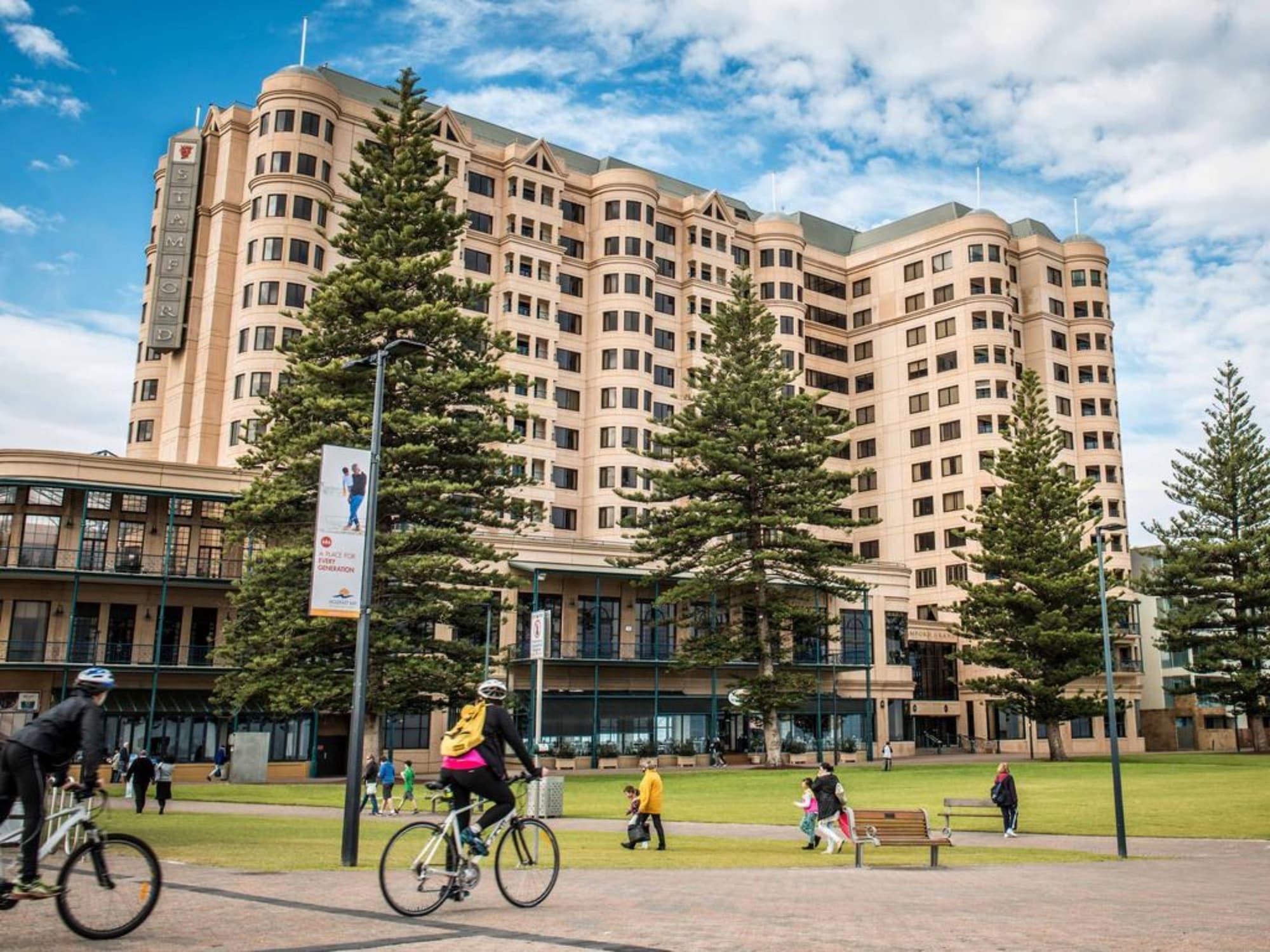 מלון גלנלג Stamford Grand Adelaide מראה חיצוני תמונה The photo depicts a modern building with a large facade, likely a hotel or commercial establishment, situated near a spacious green area. Surrounding the building are several tall trees, providing shade and enhancing the aesthetic appeal. In the fore
