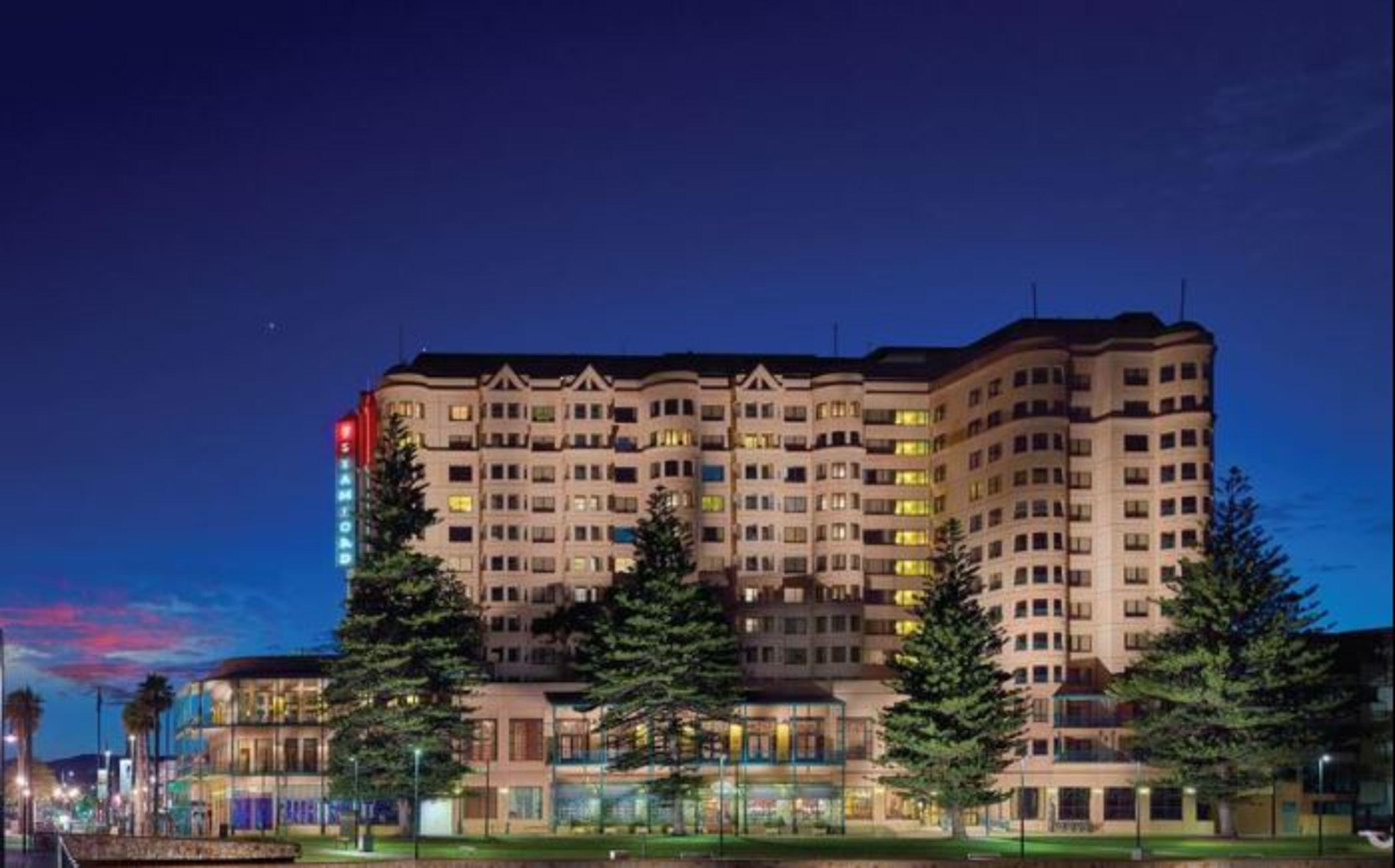 מלון גלנלג Stamford Grand Adelaide מראה חיצוני תמונה The photo displays a multi-story hotel building at dusk, illuminated by various lights. The architecture features a mix of modern and traditional design elements, with numerous windows showcasing a warm glow. Surrounding the hotel are several tall tr