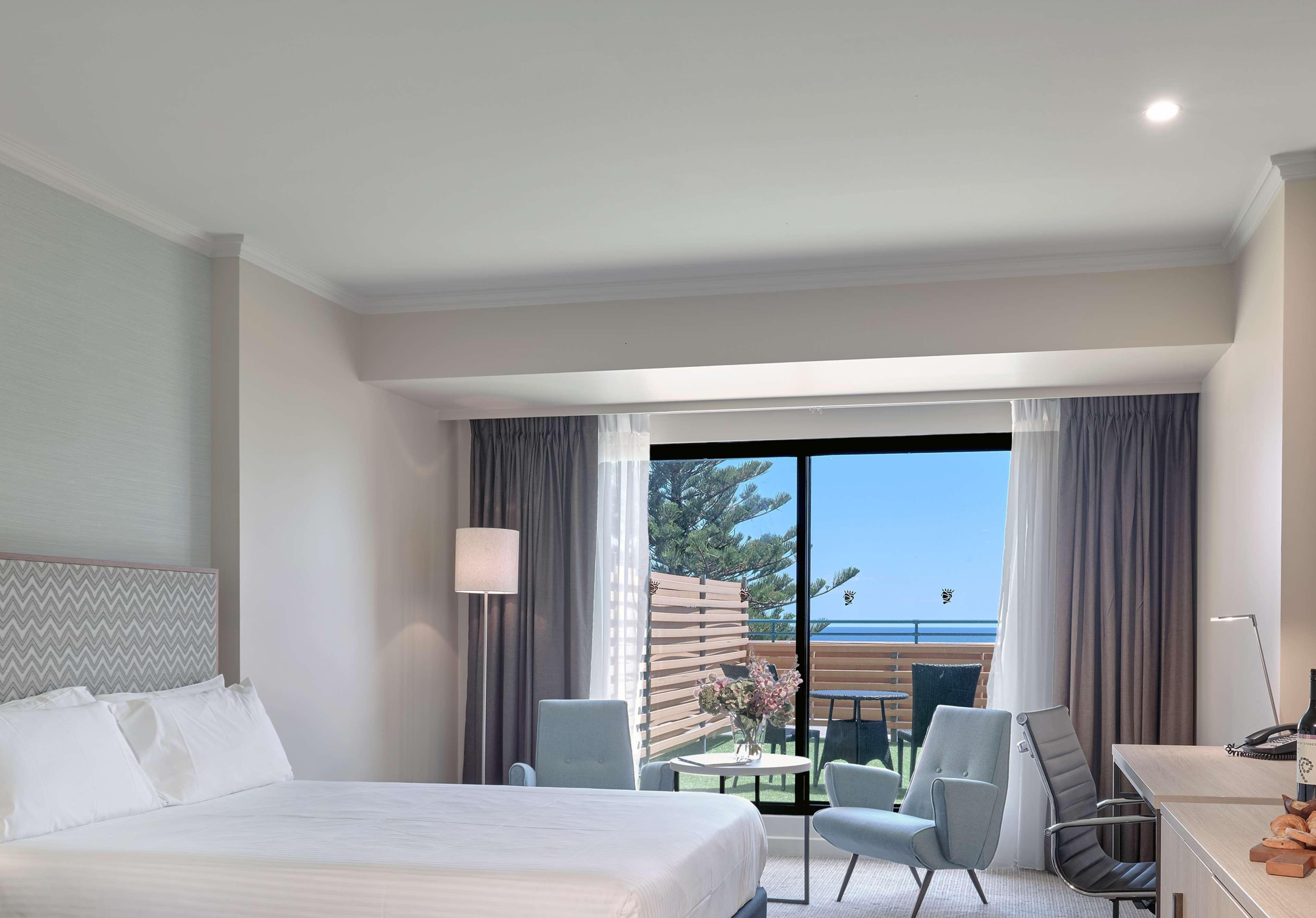 מלון גלנלג Stamford Grand Adelaide מראה חיצוני תמונה The photo shows a modern hotel room featuring a large bed with white bedding. There are two armchairs positioned near a window that offers a view of an outdoor area, possibly a balcony or patio with greenery. The room has light-colored walls and curt
