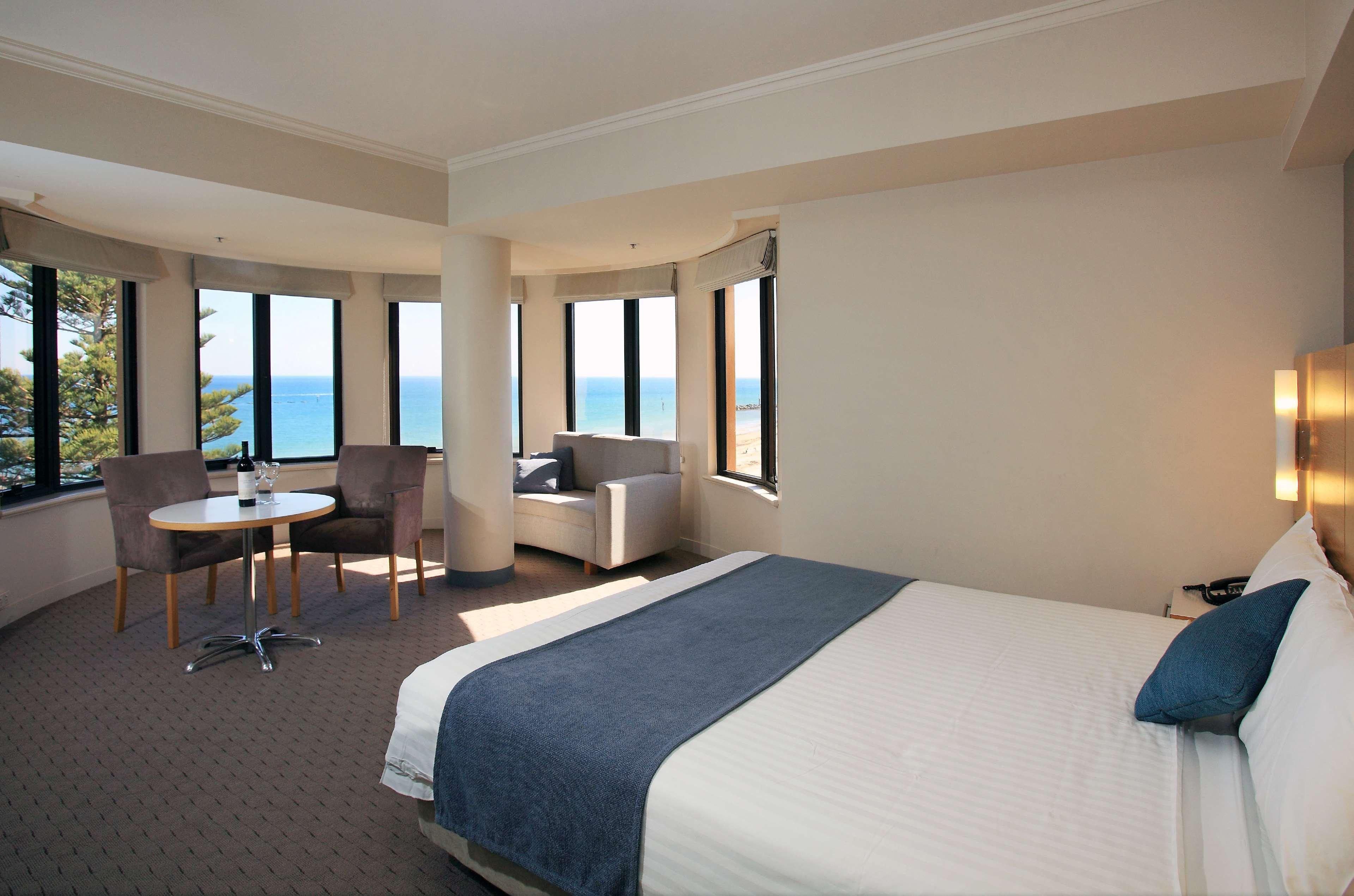 מלון גלנלג Stamford Grand Adelaide מראה חיצוני תמונה The photo shows a well-lit hotel room featuring large windows that offer a view of the ocean. The room is arranged with a comfortable bed covered in white linens and a navy blue throw blanket. There is a small table and chairs set up beside the windo