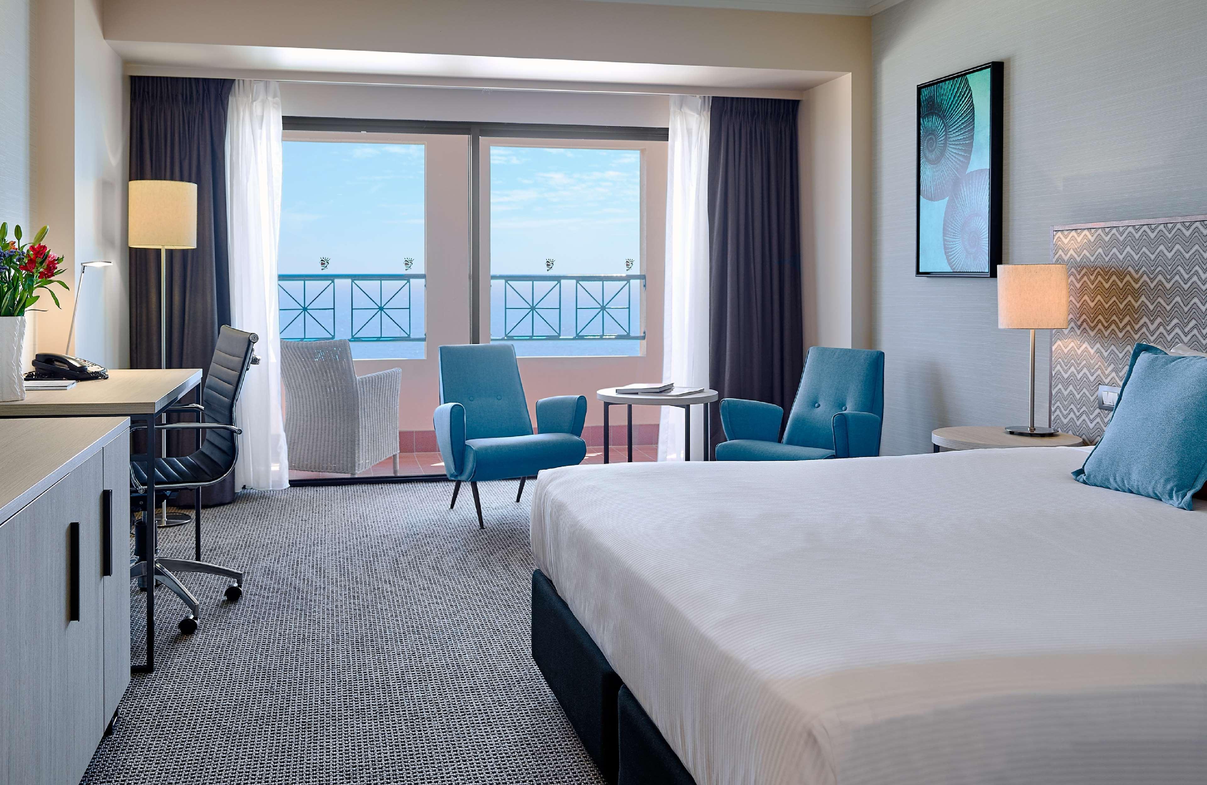 מלון גלנלג Stamford Grand Adelaide מראה חיצוני תמונה The image shows a hotel room featuring a large bed with a white duvet. There are two bright blue chairs and a small table near a large window that provides a view outside, possibly to a balcony. The room includes a workspace with a desk and an office