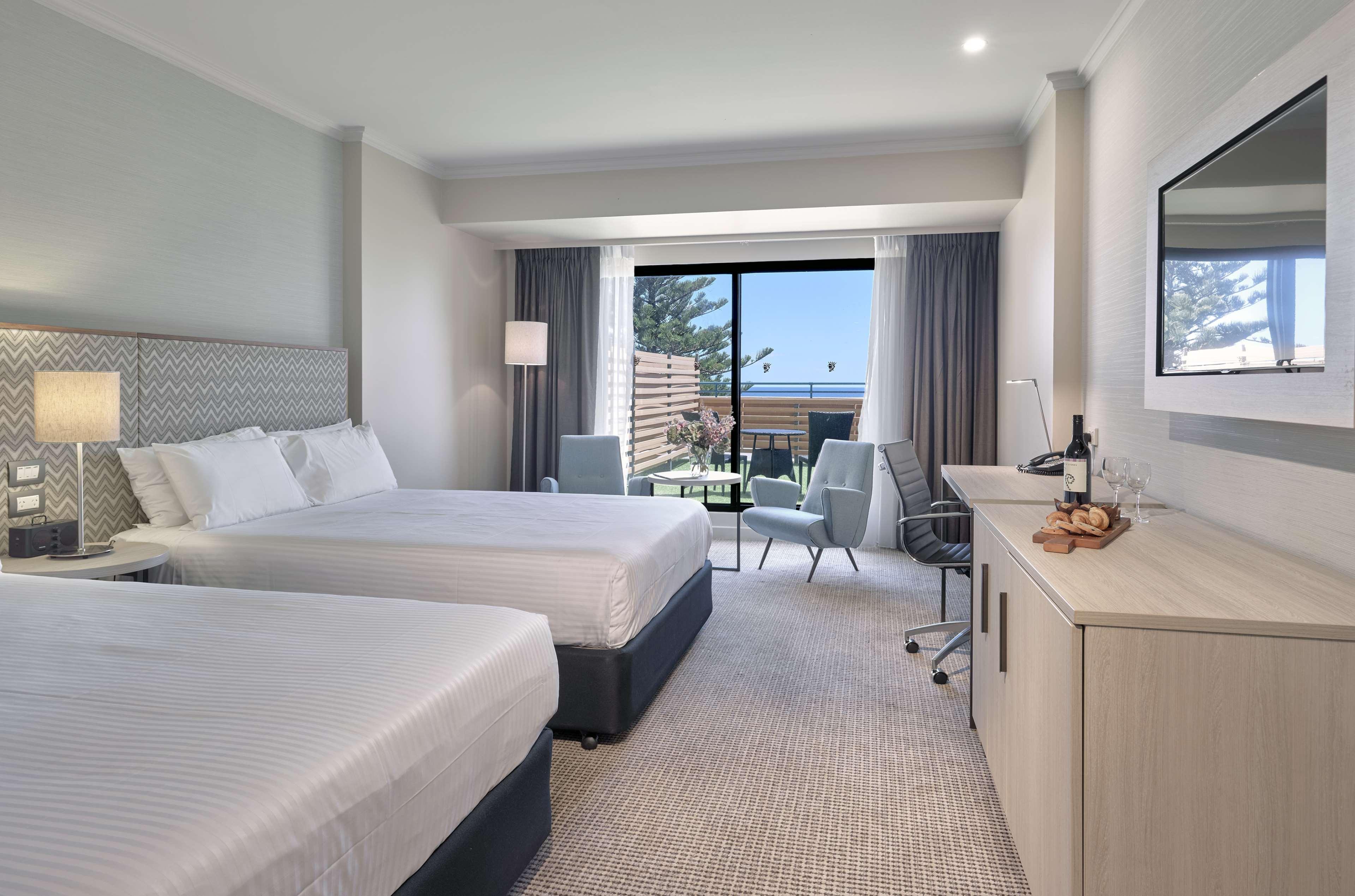 מלון גלנלג Stamford Grand Adelaide מראה חיצוני תמונה The photo shows a modern hotel room featuring two beds with white linens. There's a large window that provides natural light and a view of a balcony area. The room includes two chairs and a small table, creating a cozy sitting area. A television is m