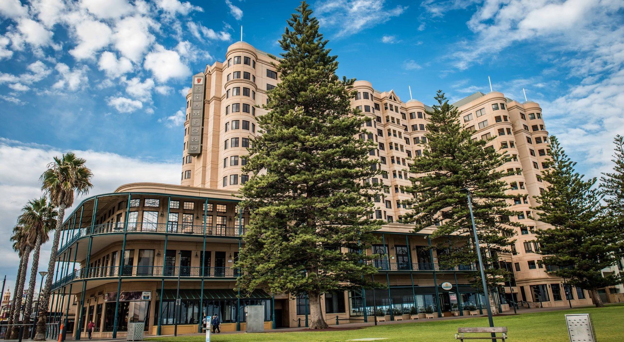 מלון גלנלג Stamford Grand Adelaide מראה חיצוני תמונה The image shows a large building with a distinctive architectural style, characterized by a tan or beige color. The structure appears to have multiple floors and rounded edges. In front of the building, there are several tall green trees, which add t