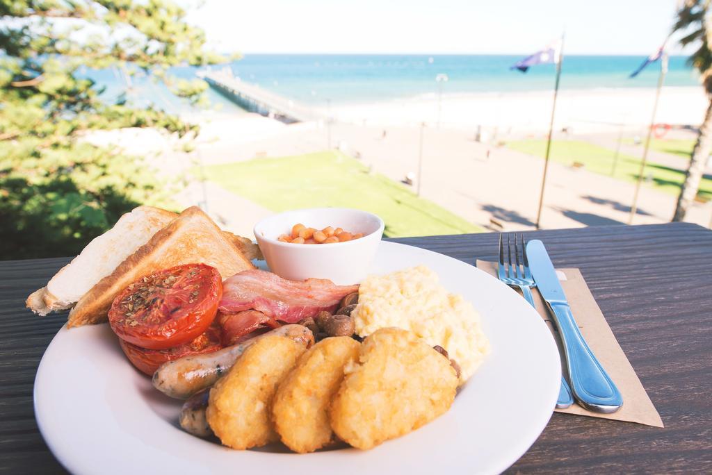 מלון גלנלג Stamford Grand Adelaide מראה חיצוני תמונה The photo shows a plate of breakfast food set against a beachside backdrop. The plate includes several items: crispy bacon, a grilled tomato, sausage links, golden hash browns, and a small serving of baked beans. There is also a piece of toast on the